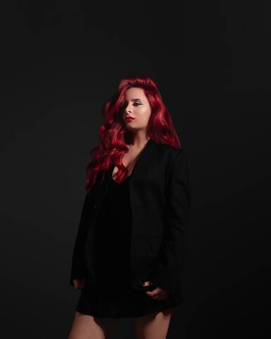 CERES posing against a dark background, wearing a black outfit with long red hair cascading over her shoulders in a strong, editorial portrait.