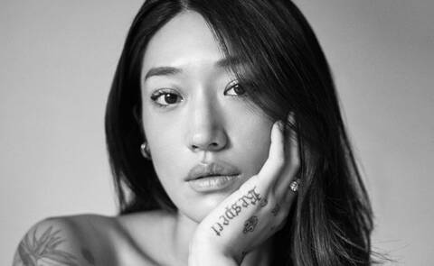 Black and white portrait of Peggy Gou looking at the camera with her face resting on one hand, with tattoos visible on her hand and arm.