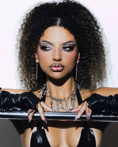 Studio portrait of Clementaum wearing bold graphic makeup, silver jewelry, and black gloves, posing with both hands on a metal rail against a light background.