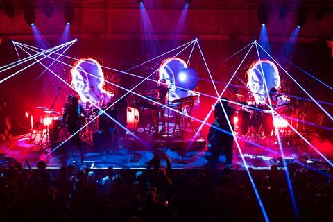 Faithless performing live on stage with red lighting, laser beams, and large digital visuals behind the band, seen from the audience perspective.