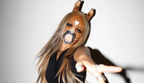 horsegiirl posing with horse inspired face makeup and long blonde hair, extending one arm toward the camera against a plain background.