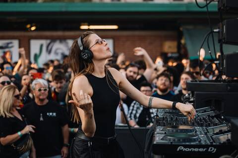Charlotte de Witte performing at an outdoor electronic music event, wearing headphones and sunglasses while mixing on Pioneer DJ equipment in front of a large crowd.